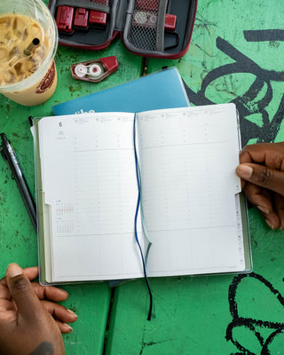 An open weekly planner with two ribbon bookmarks, held by two hands over a green table. A blue notebook, a black pen, a red tape dispenser, and an iced coffee are visible around it.