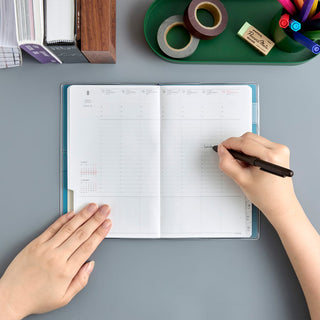 An open weekly planner with a person writing, shown from above on a gray desk. A tray with washi tape, markers, and a pencil is visible at the top, along with books on the left.