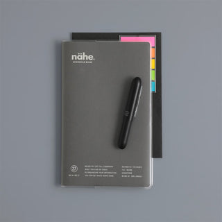 A gray schedule book with the brand name 'nähe.' printed on the cover, accompanied by a black pen and a black folder with colorful tabs, arranged on a solid gray background.