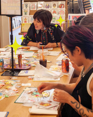 People drawing at a craft table with pens, papers, and paint jars