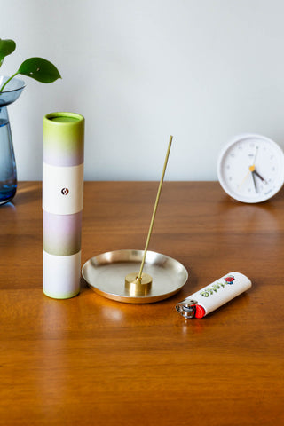 A small round brass tray with matching brass incense holder holds a light green stick of compressed powder incense. A lighter sits beside it on one side, and the incense box on the other. The incense box is a pretty green to purple gradient with gold emboss details.