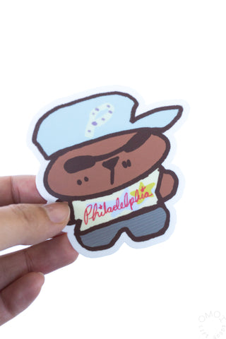 Hand holding a bear sticker with blue hat and Philadelphia shirt on a white background