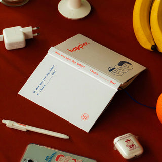 Notebook opened with a pen on a red surface, featuring the name 'happier'. and cartoons on the cover