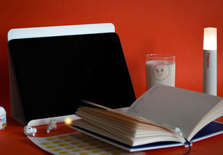 Tablet on a desk with a notebook, glass of milk, and lamp against an orange wall.