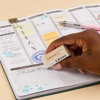 Hand using a small eraser on a planner with a pen, on a beige surface.